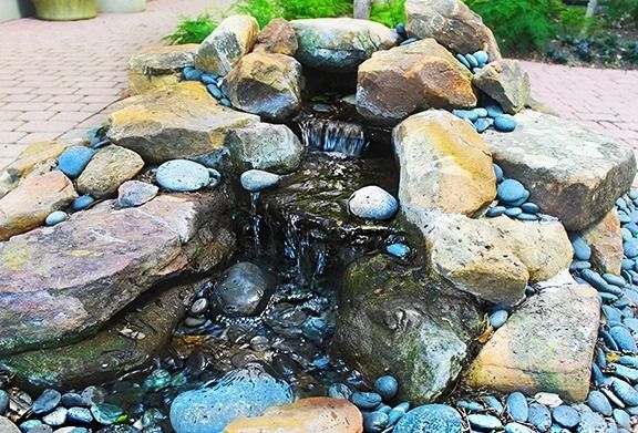 Small rock waterfall cascading into a pool. Brown and grey rocks are arranged around the water.