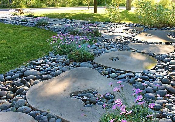 Stepping stones and black river rocks create a garden path. Pink flowers bloom, with green grass in the background.