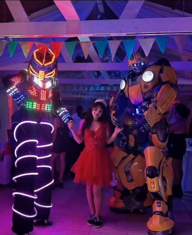 Mujer con vestido rojo posando con dos robots disfrazados. Ambiente de fiesta con luces.