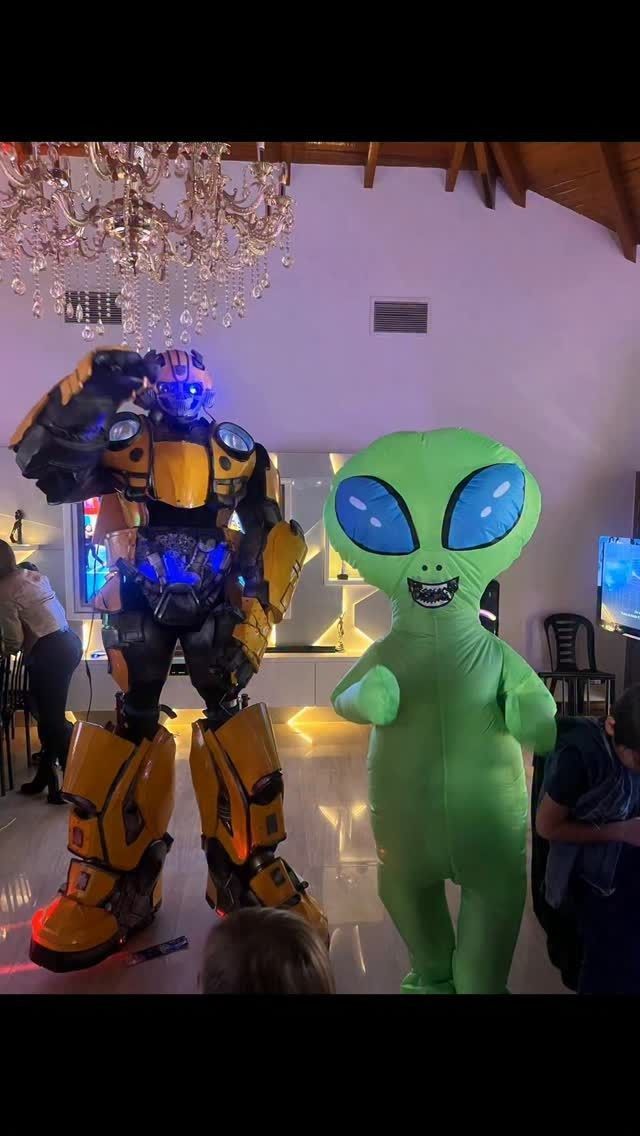 En el interior se ven disfraces de Bumblebee Transformer y de alienígenas verdes, posiblemente en una fiesta con luces cenitales.