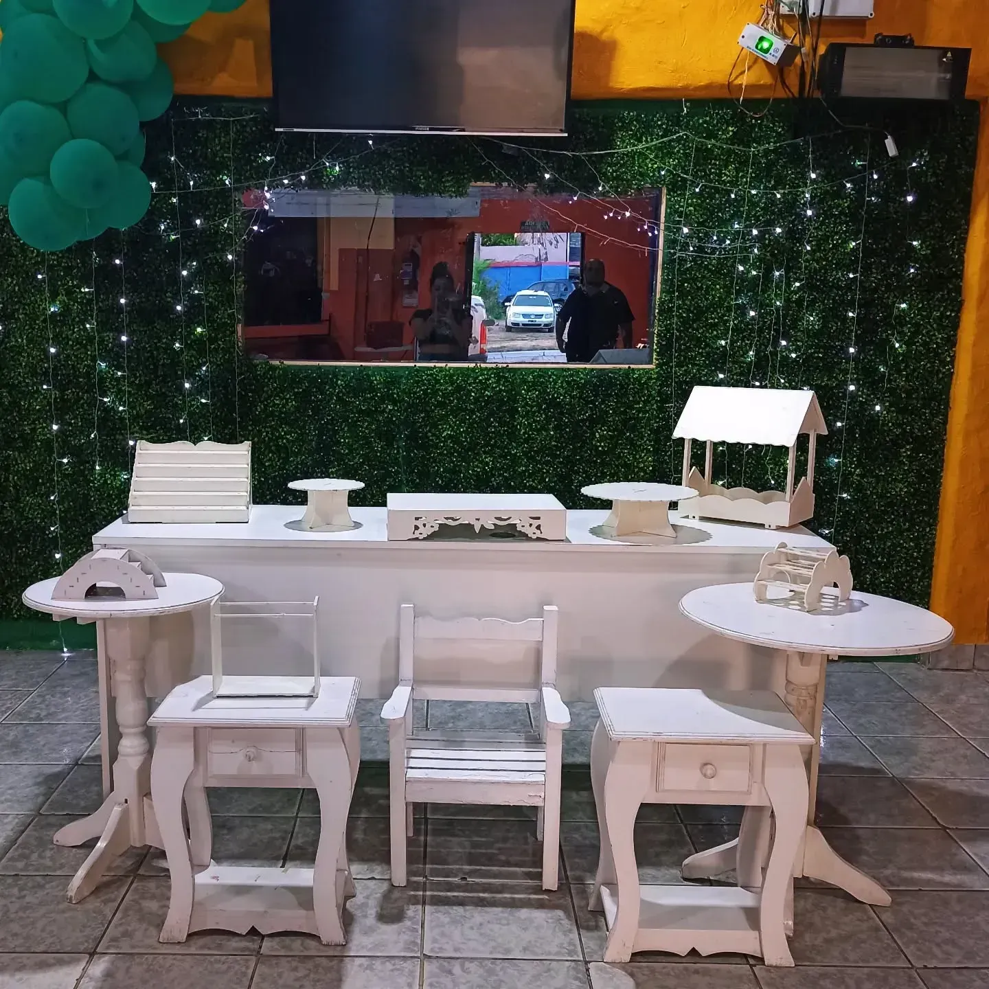 Una mesa de postres blanca con decoración a juego, con un fondo de follaje verde, luces de hadas y un televisor.