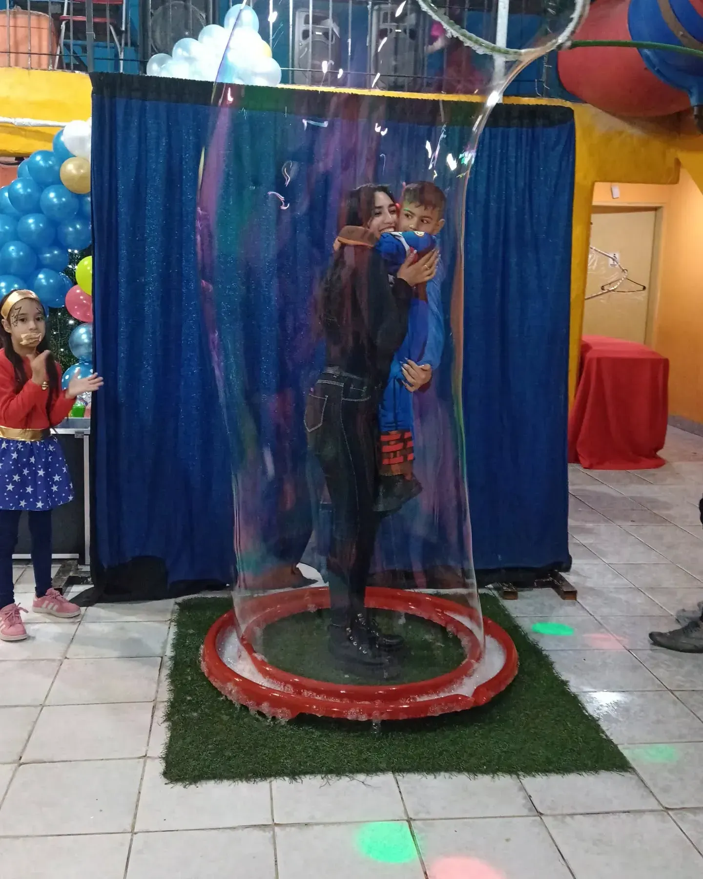 Mujer sosteniendo a un niño dentro de una burbuja gigante en una fiesta, fondo azul, otro niño observa.