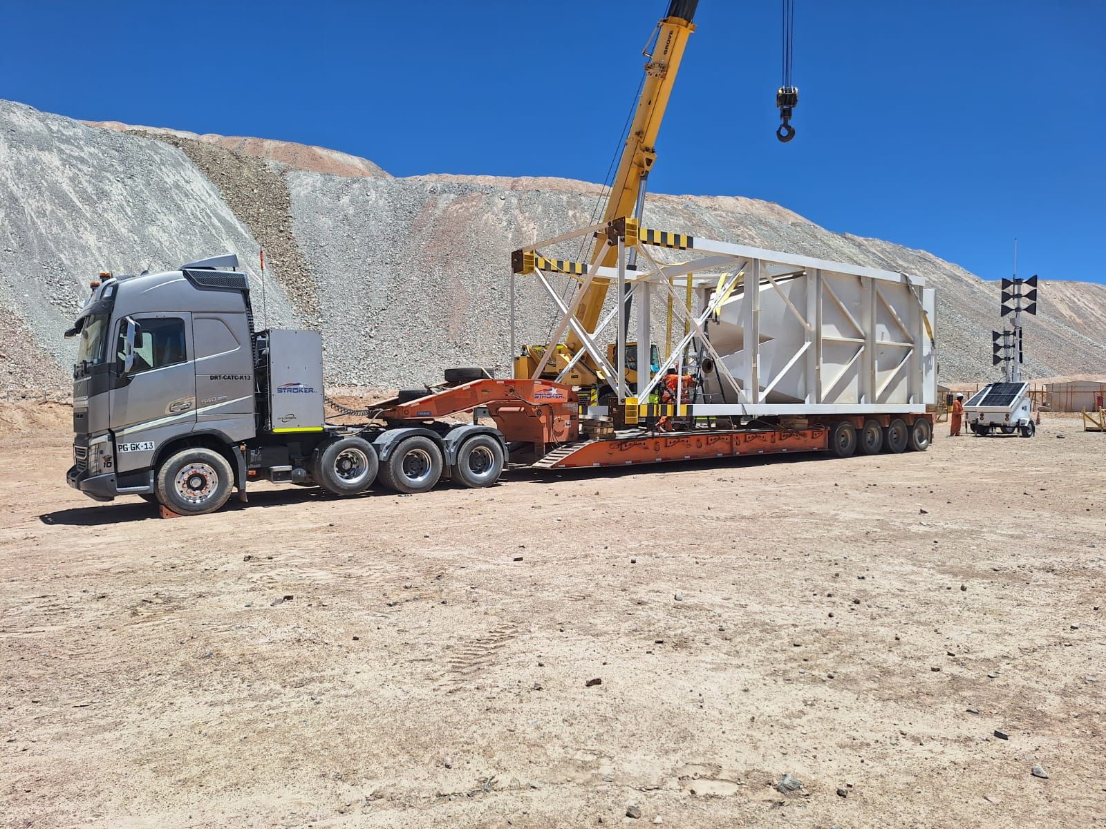 Excavadora Komatsu amarilla en un remolque de plataforma, con señal de precaución, bajo un cielo azul.
