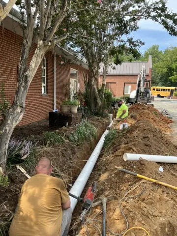 Construction workers install white pipe in a trench next to a brick building. Yellow safety vests, and an excavator are visible.