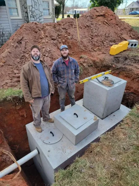 Two men stand near a newly installed concrete septic tank in a residential yard with dirt mound.