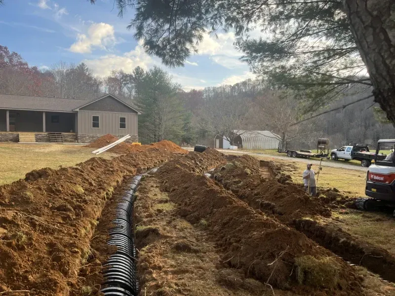 A trench dug in a yard with black drain pipe. Workers and machinery near a house in a rural setting.