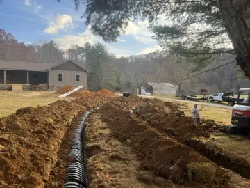 A trench dug in a yard with black drain pipe. Workers and machinery near a house in a rural setting.