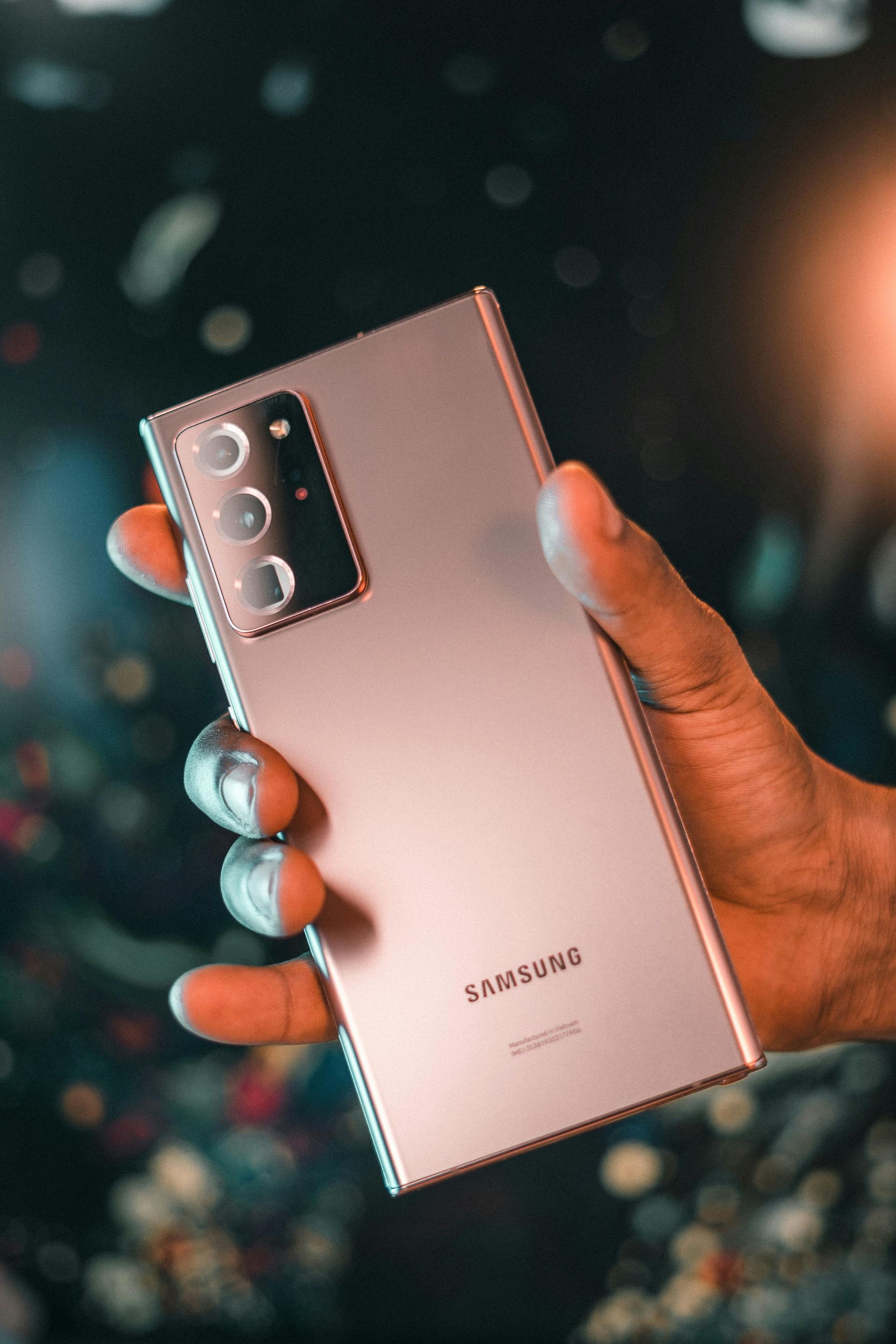Mano sosteniendo un Samsung Galaxy Note20 de oro rosa con lente de cámara triple, fondo borroso.