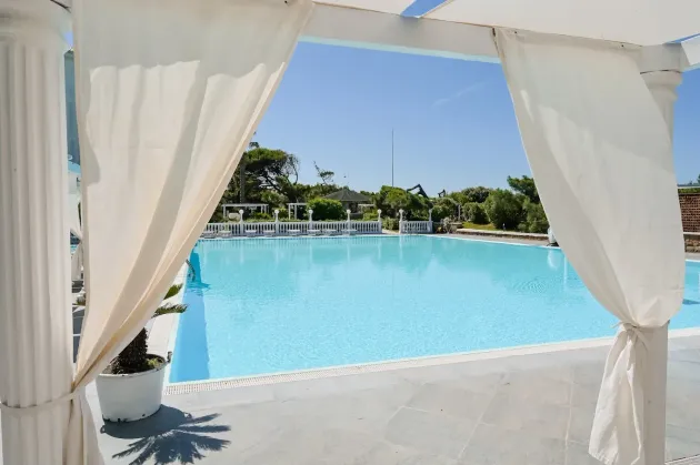 Tende bianche incorniciano una piscina turchese brillante, con lettini prendisole e alberi sullo sfondo.