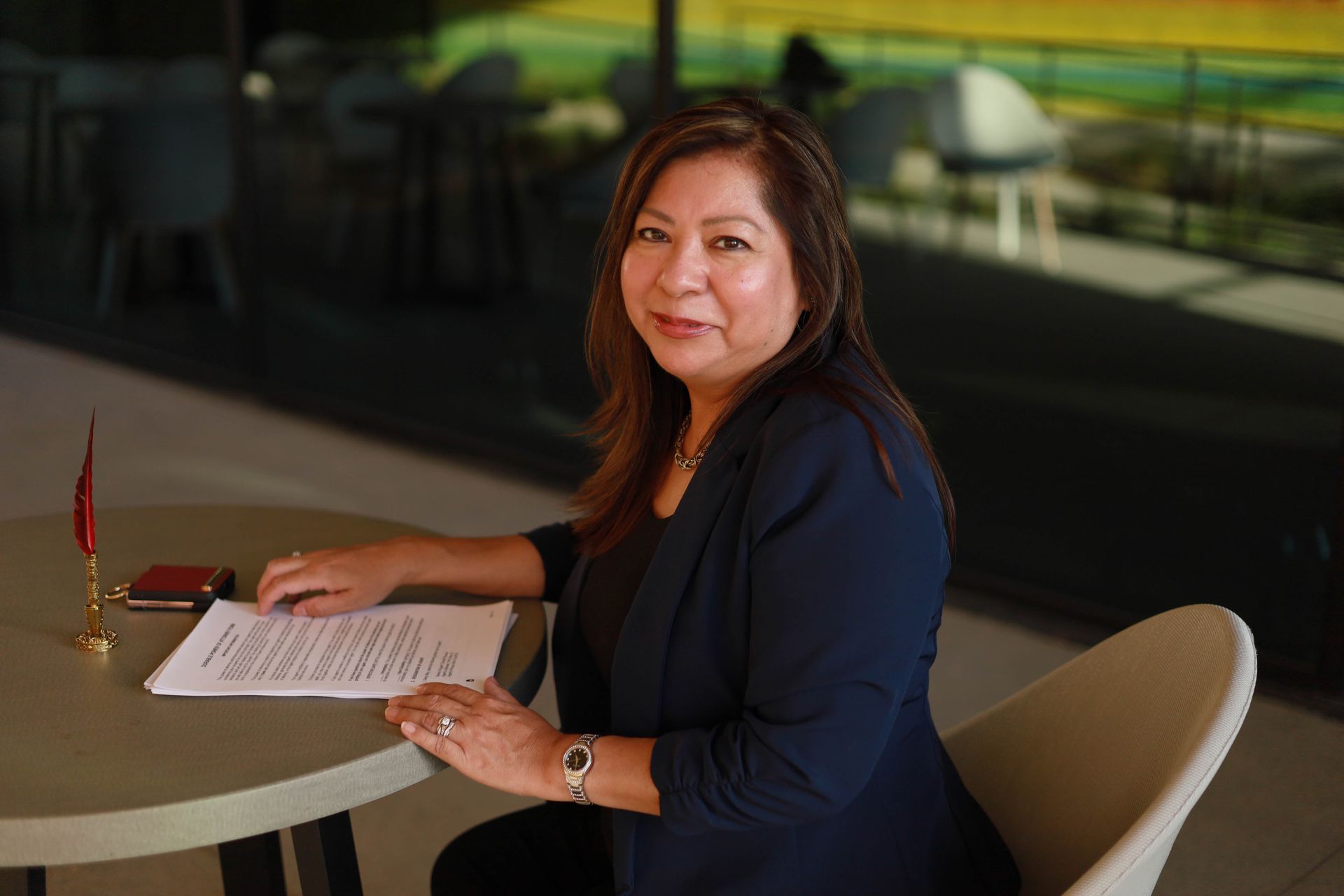 Woman in a dark blazer sits at a table, reviewing papers, in a modern setting with natural light.