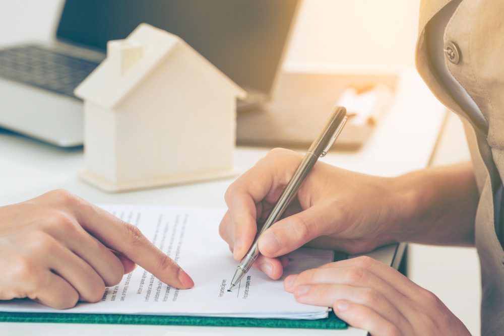 Person signing real estate contract; hand pointing; house model and laptop in background.