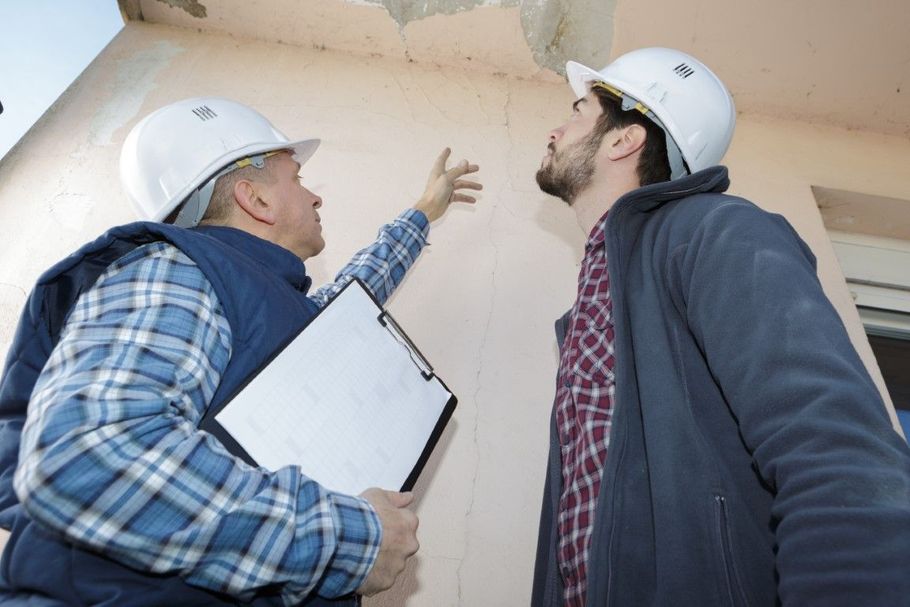 Two workers in hard hats inspecting a ceiling, one pointing while holding a clipboard in a building interior