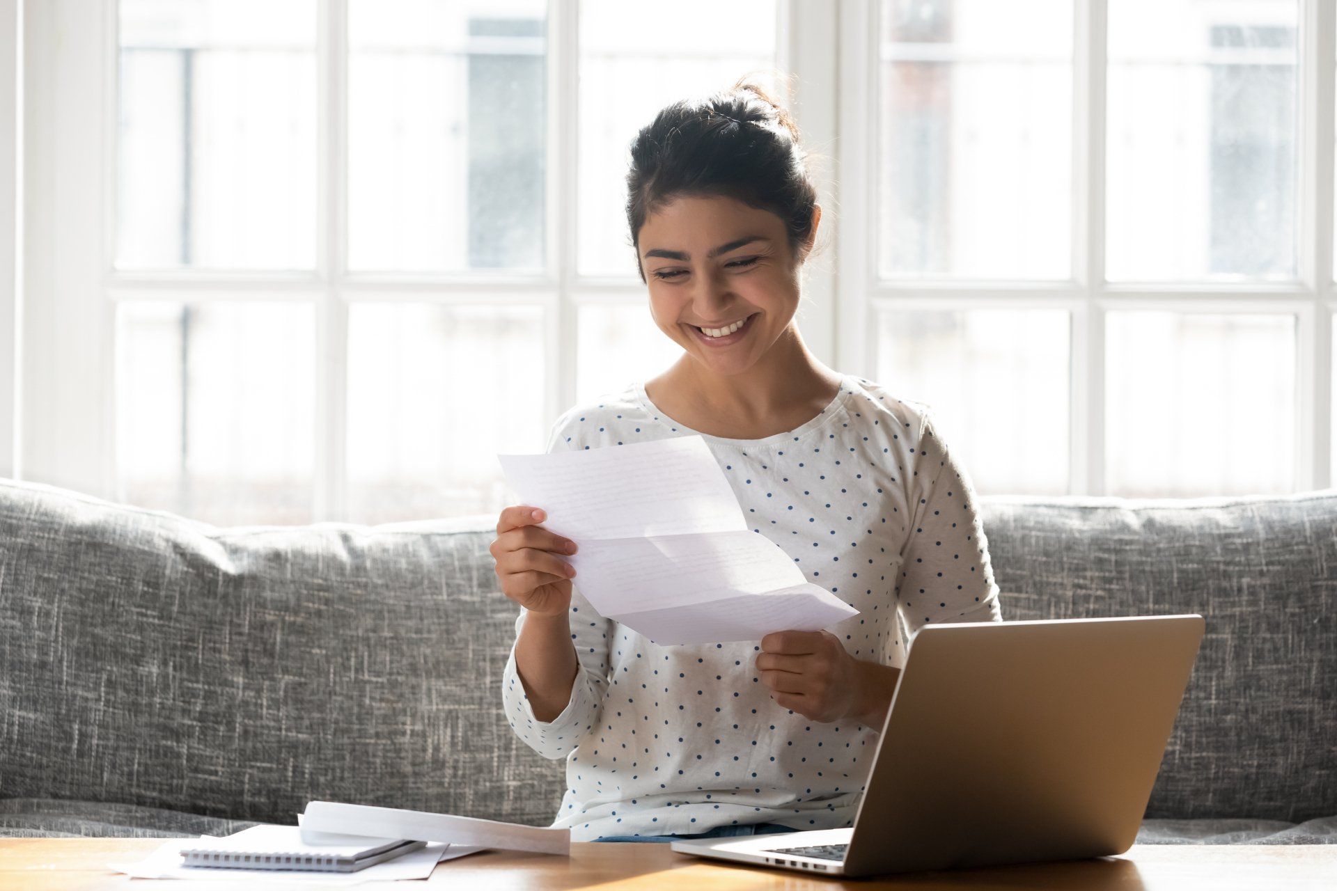 Indian ethnicity woman sitting on couch at home reading paper notice receive good news feels happy, cheerful student female looking at document enjoy exam results or college admission letter concept
