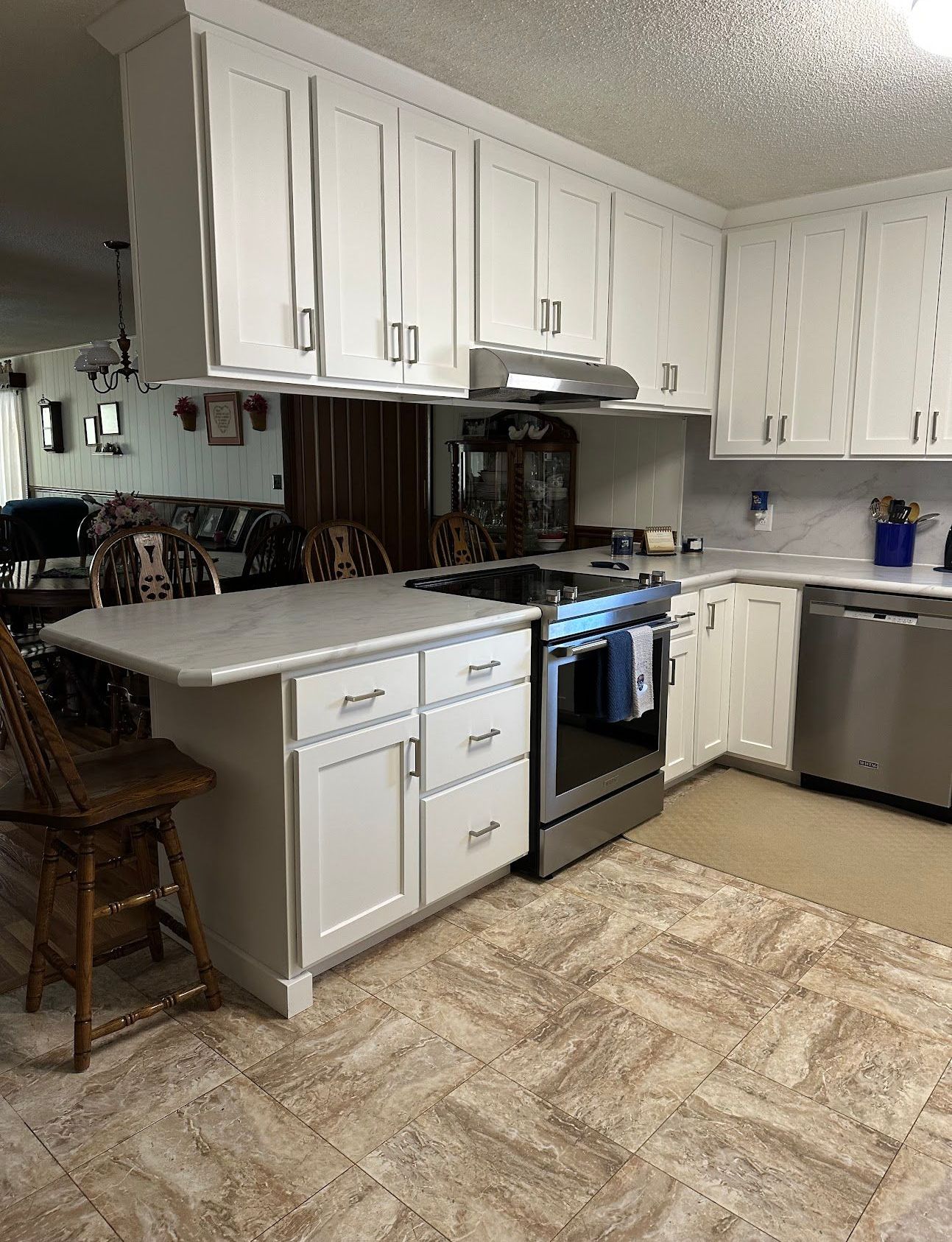 A kitchen with white cabinets , a stove , a dishwasher , and a chair.