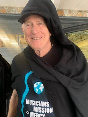 A man wearing a black shirt that says musicians mission of mercy is standing in front of a car.