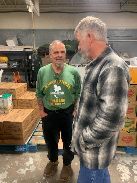 Two men are standing next to each other in a warehouse talking.