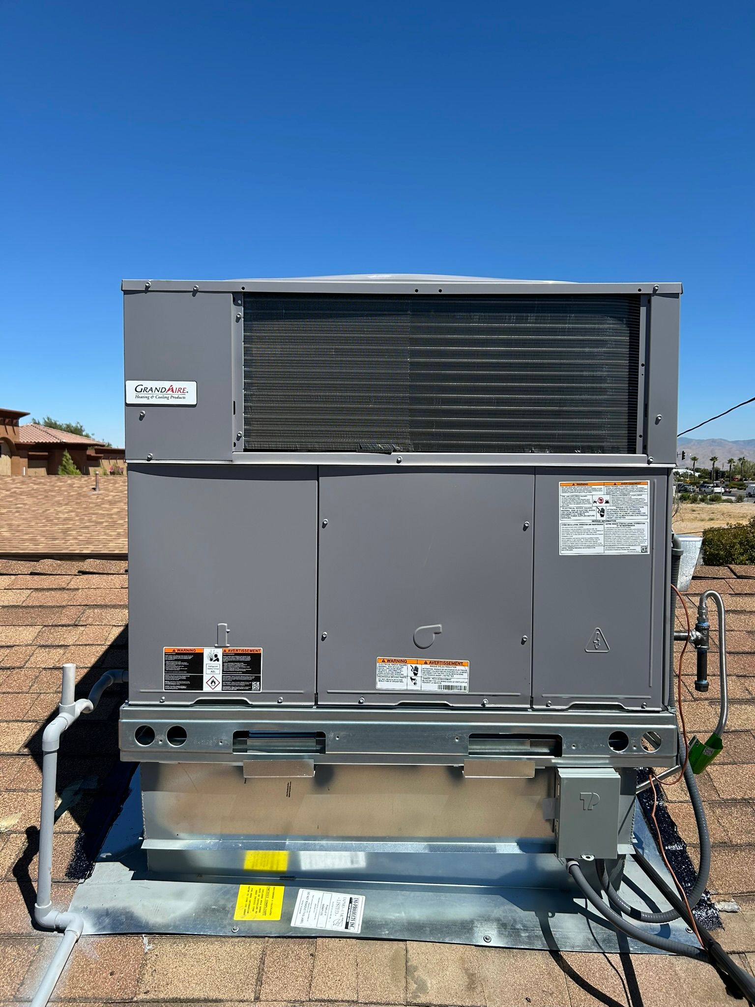 A rooftop HVAC unit, gray and silver, sits on brown shingles under a clear blue sky.