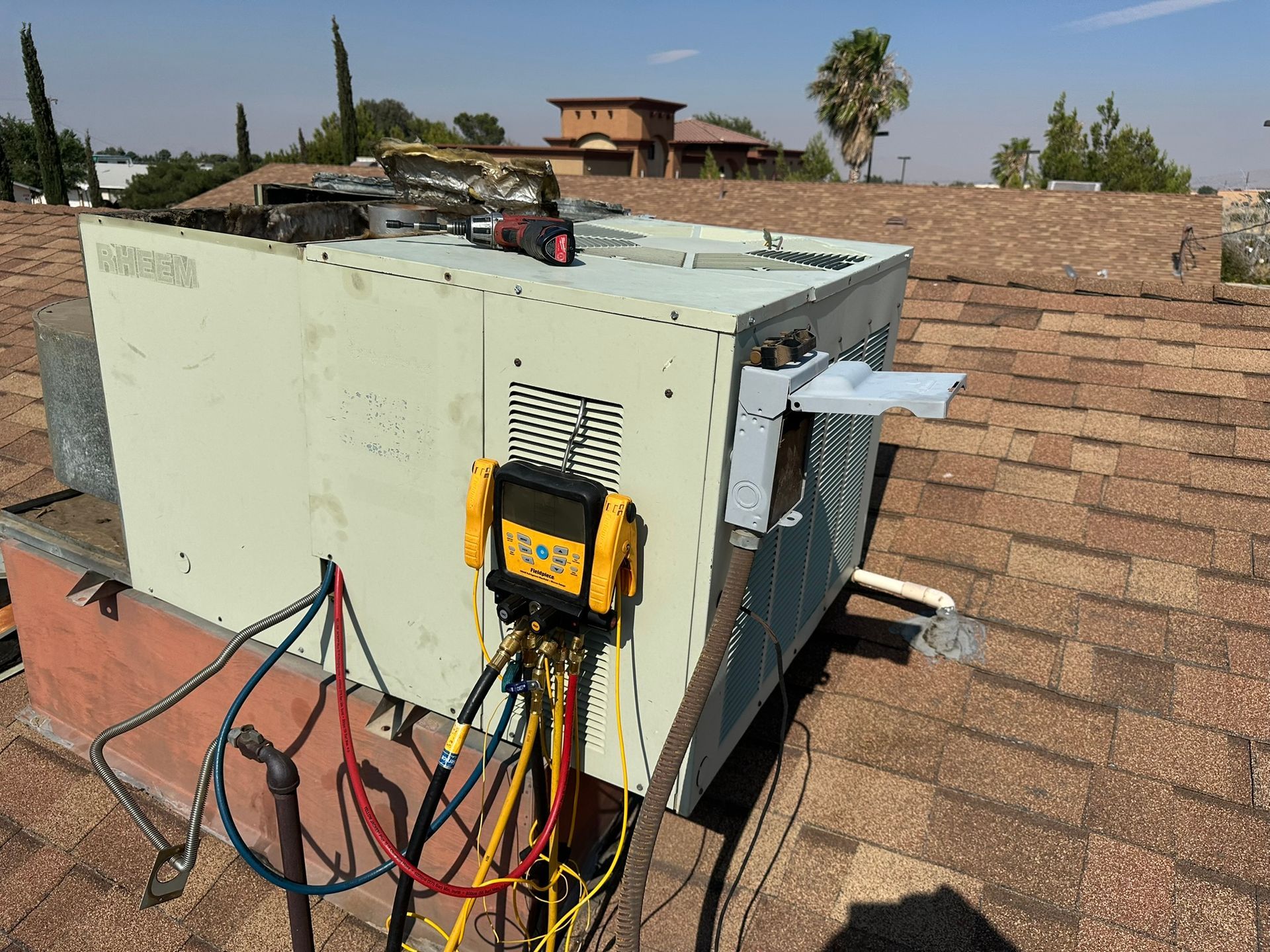 HVAC unit on a rooftop with attached gauges and tubing. Beige metal box against brown roof tiles, sunny sky.
