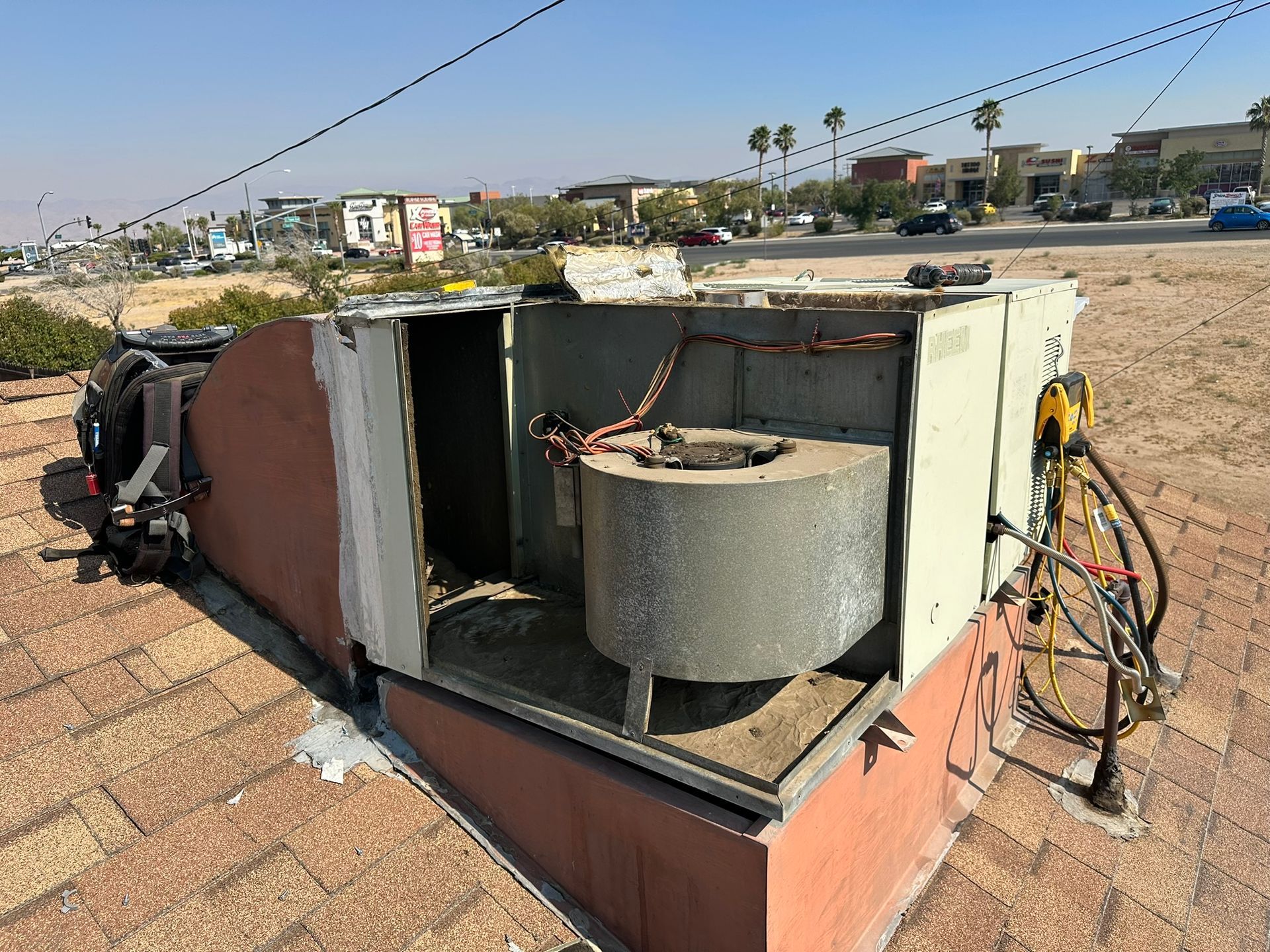 An open air conditioning unit sits on a brown rooftop, wires visible, with a suburban backdrop.