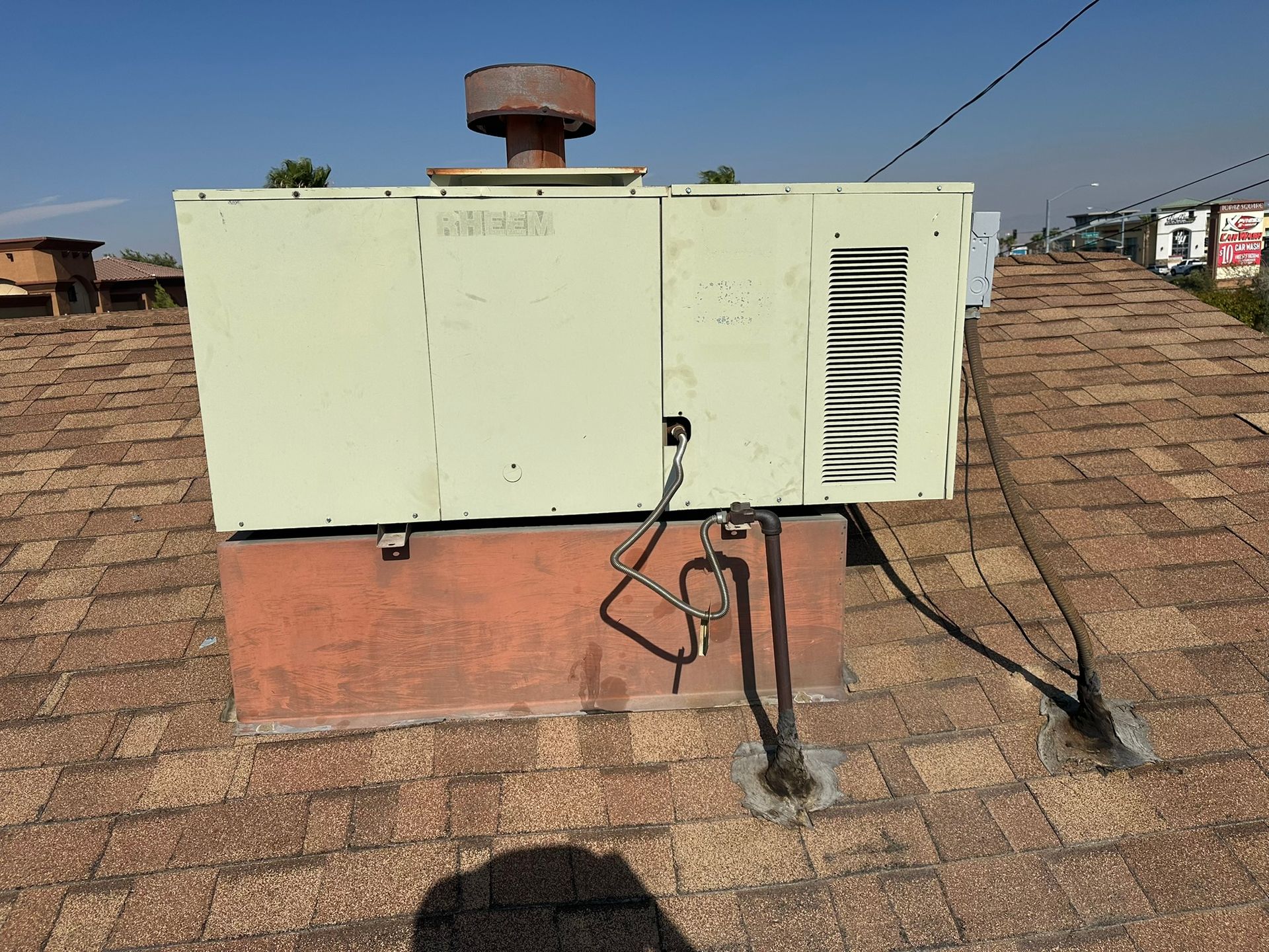 HVAC unit on a brown shingled roof, beige and rust colors, with vent and power lines.