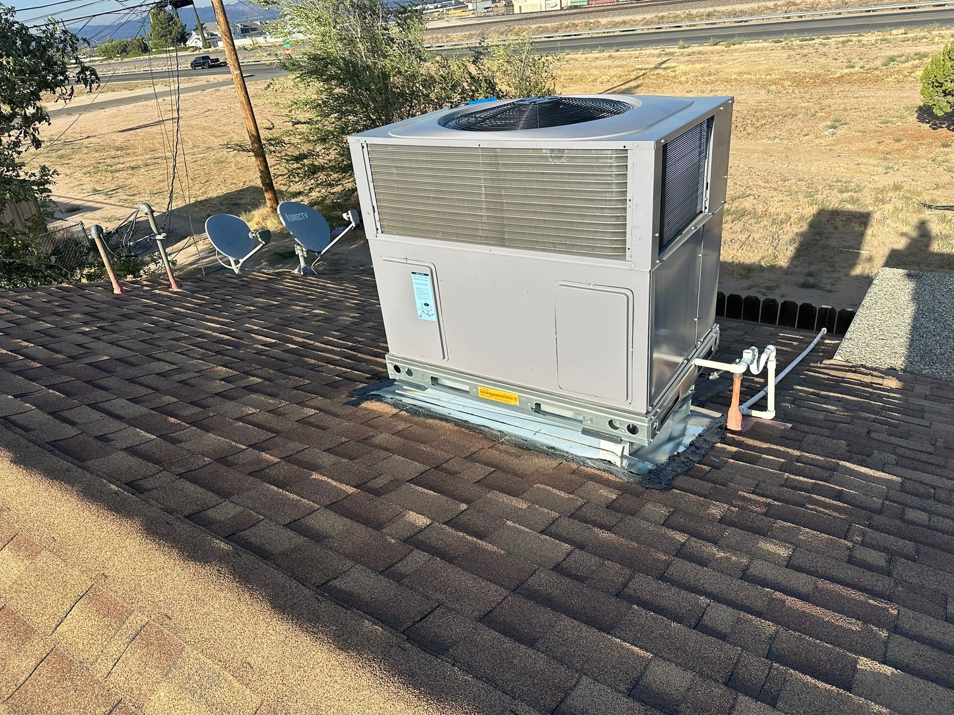 Rooftop air conditioning unit on a brown shingled roof, with satellite dishes and a ladder in the background.