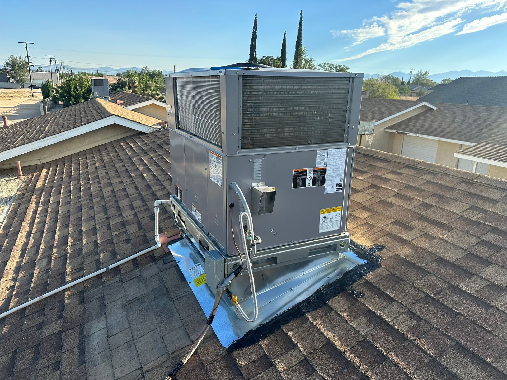 Rooftop HVAC unit on a brown shingle roof, under a blue sky, with surrounding houses and trees in the background.