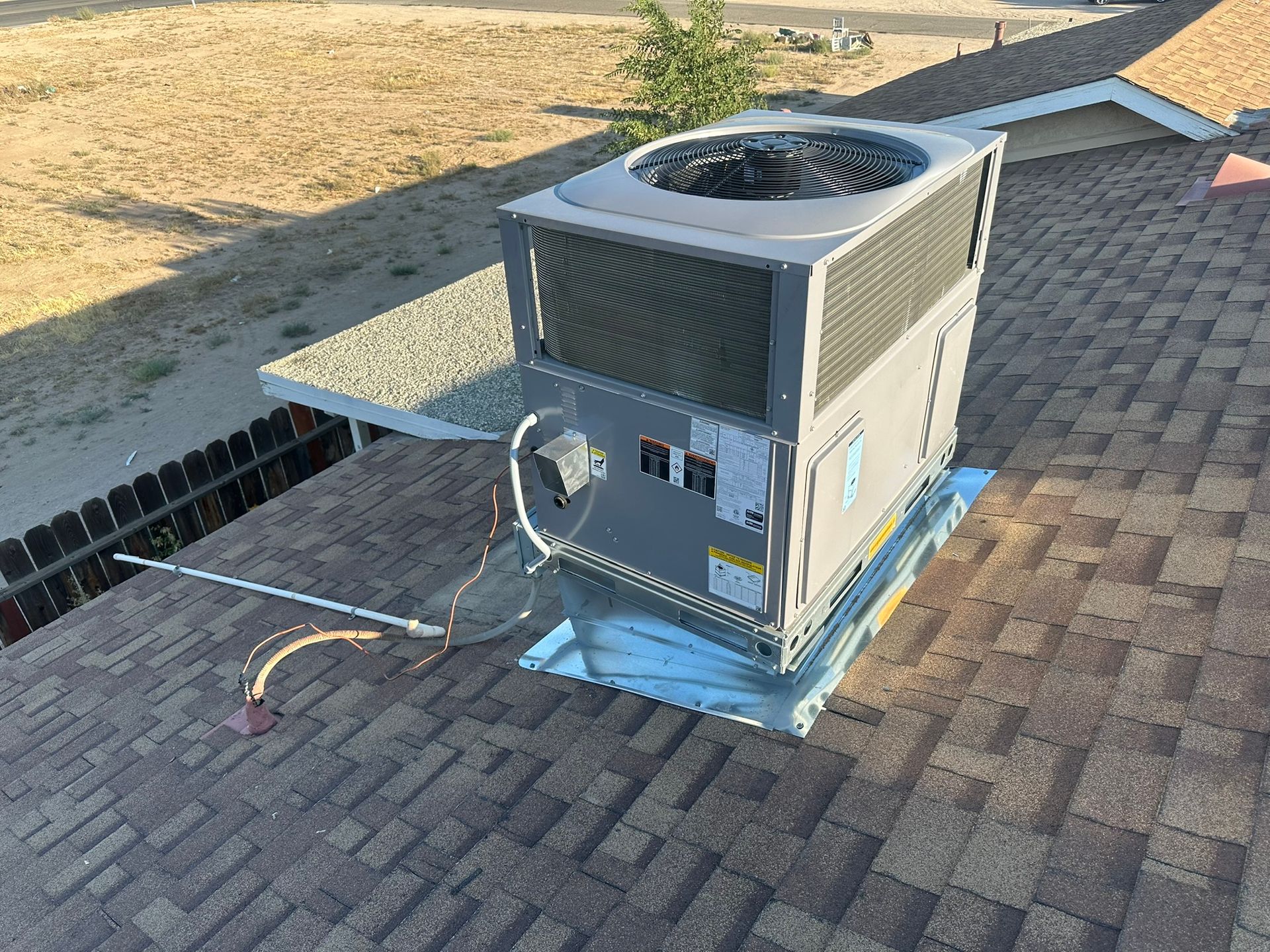 AC unit on a rooftop with brown shingles. Silver metal base, exhaust vent, and pipes.