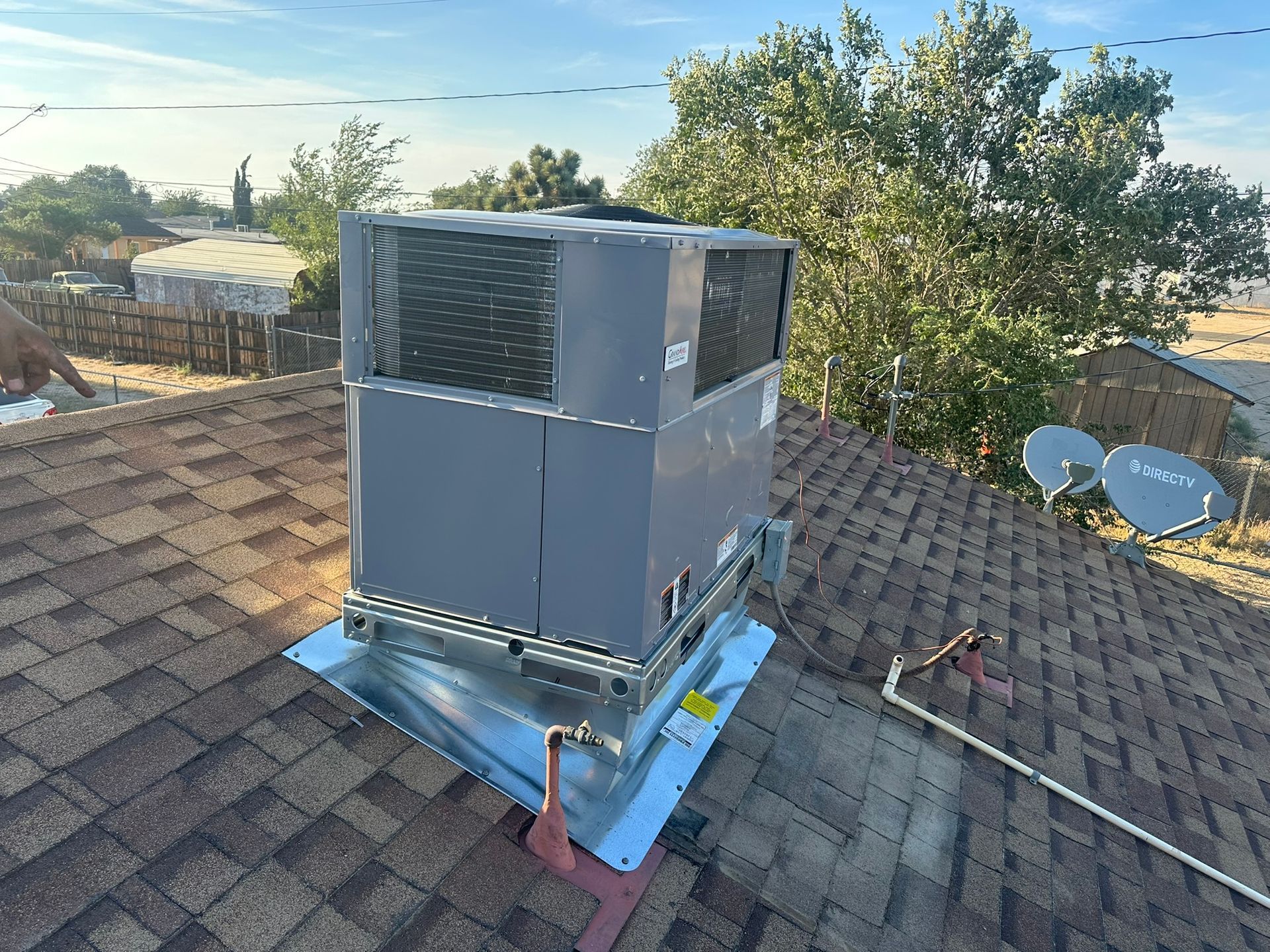 HVAC unit on a rooftop, gray and square, with satellite dishes and trees in the background.
