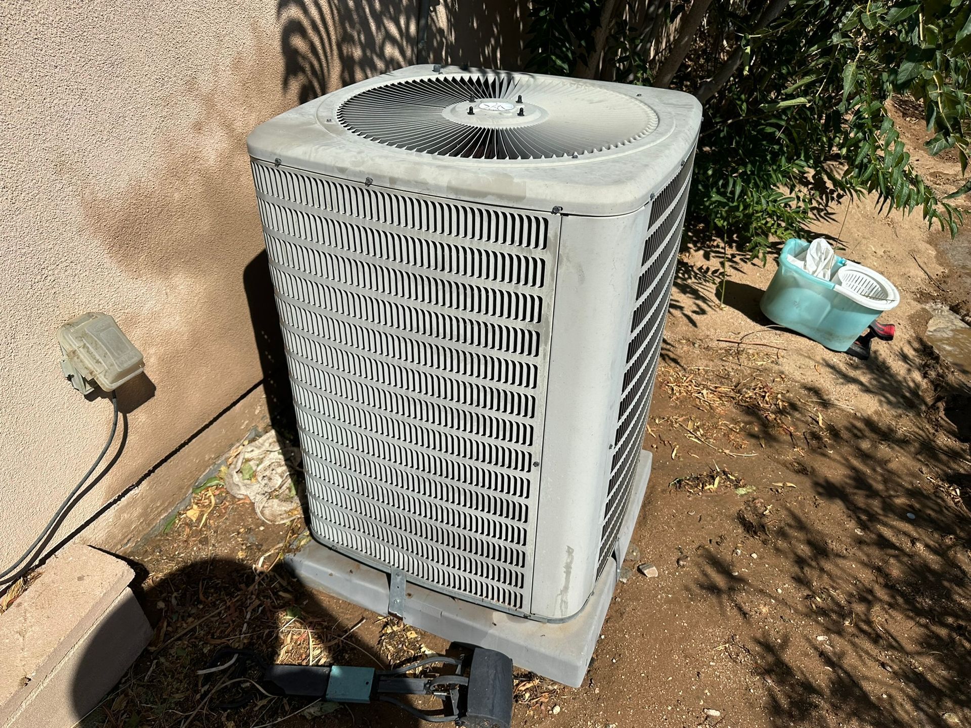 Outdoor air conditioning unit with metal grilles and fan. Grey and white, concrete base, near wall and foliage.