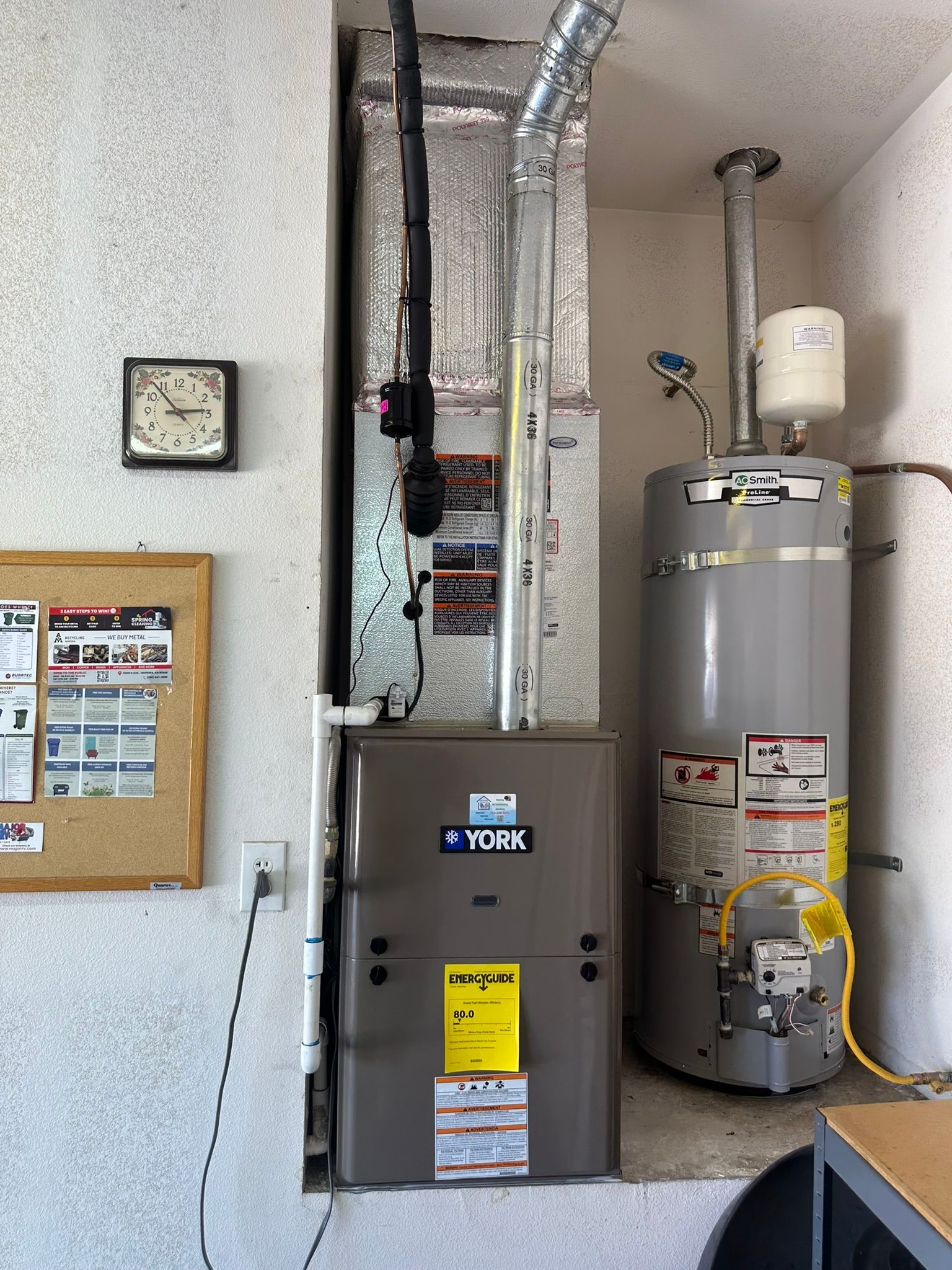 Furnace and water heater in a utility room. Gray appliances with ductwork against a textured white wall.