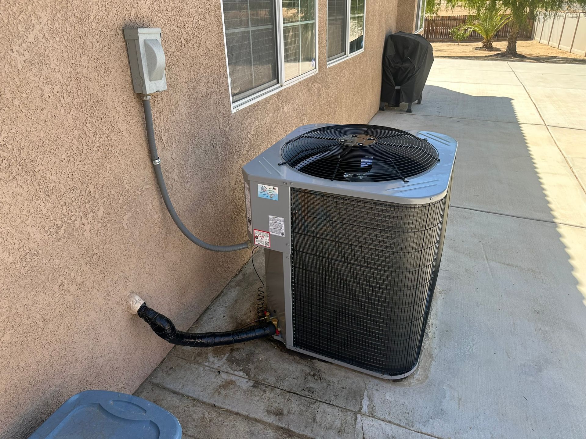 Outdoor air conditioning unit next to a stucco wall with a gray electrical box and a black hose.
