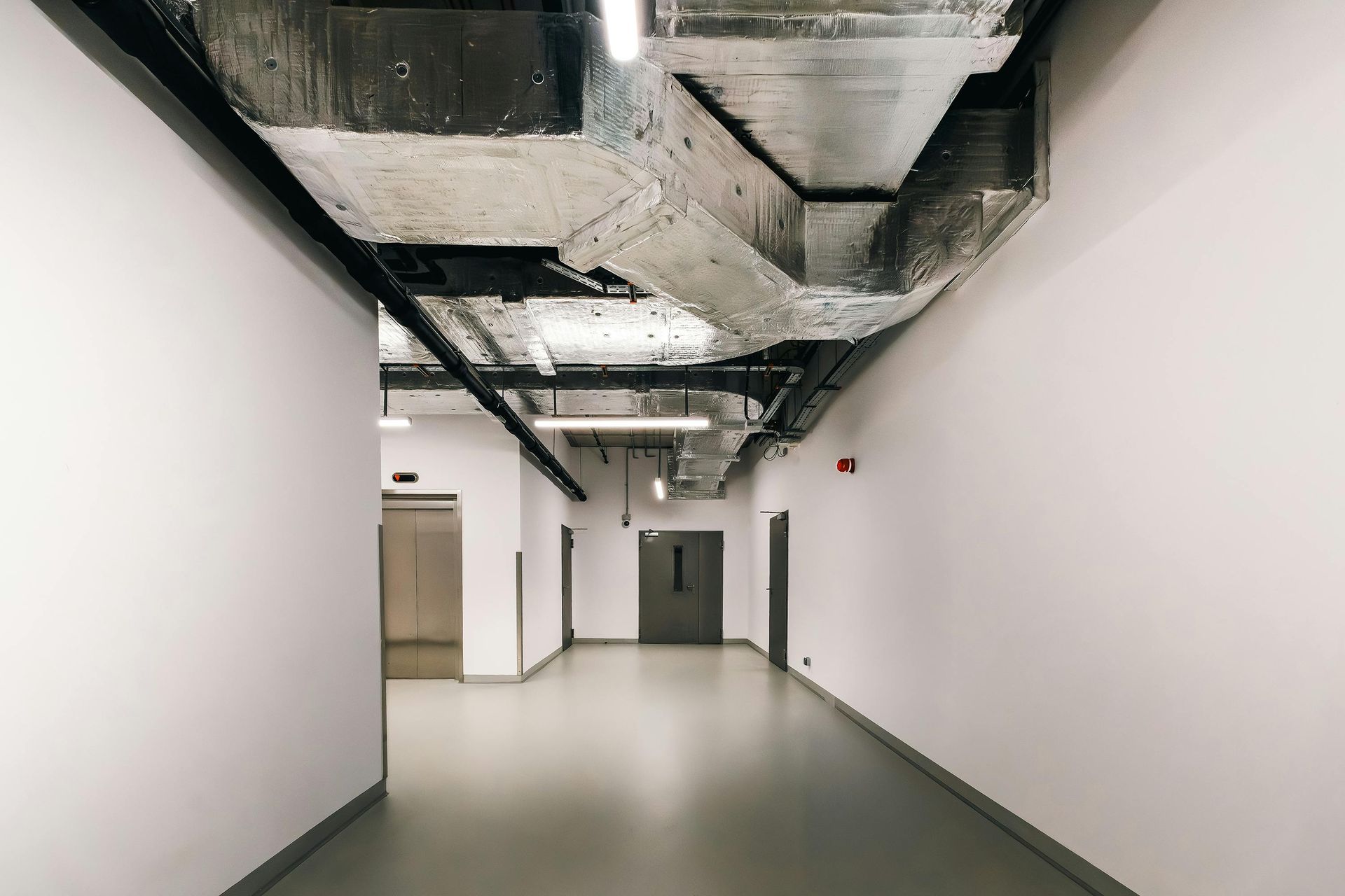 Long hallway with white walls and exposed ceiling ducts. Elevator on the left, door at the end.