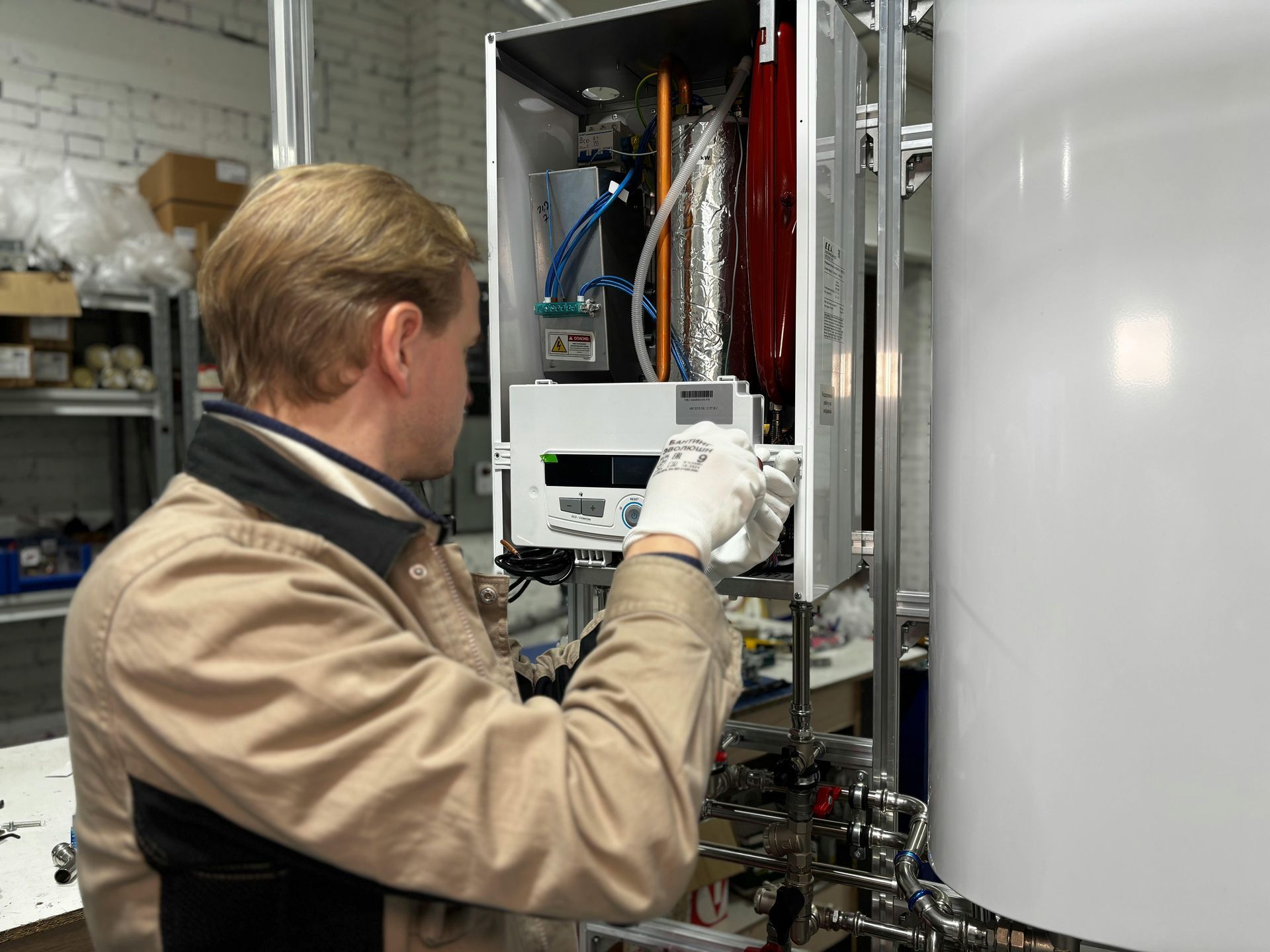 Man in tan coat assembling appliance, wearing gloves, in workshop setting.