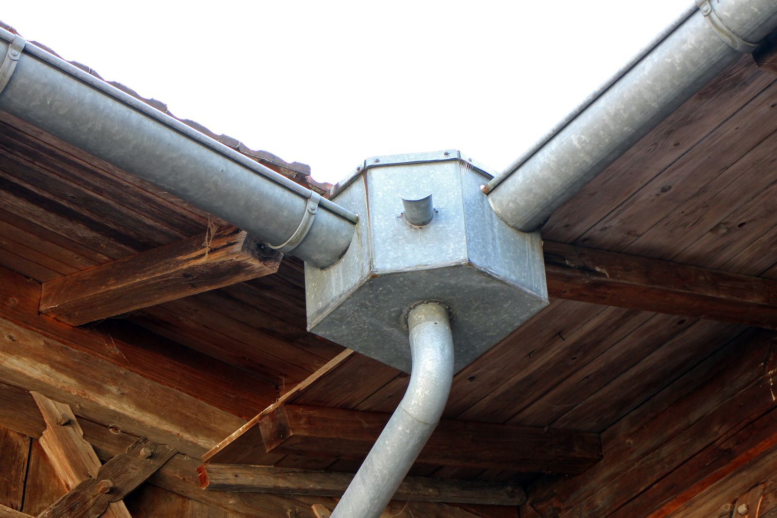 Gray metal rain gutter system with a hexagonal joint connecting two roof gutters and a downspout, attached to a wooden eave.