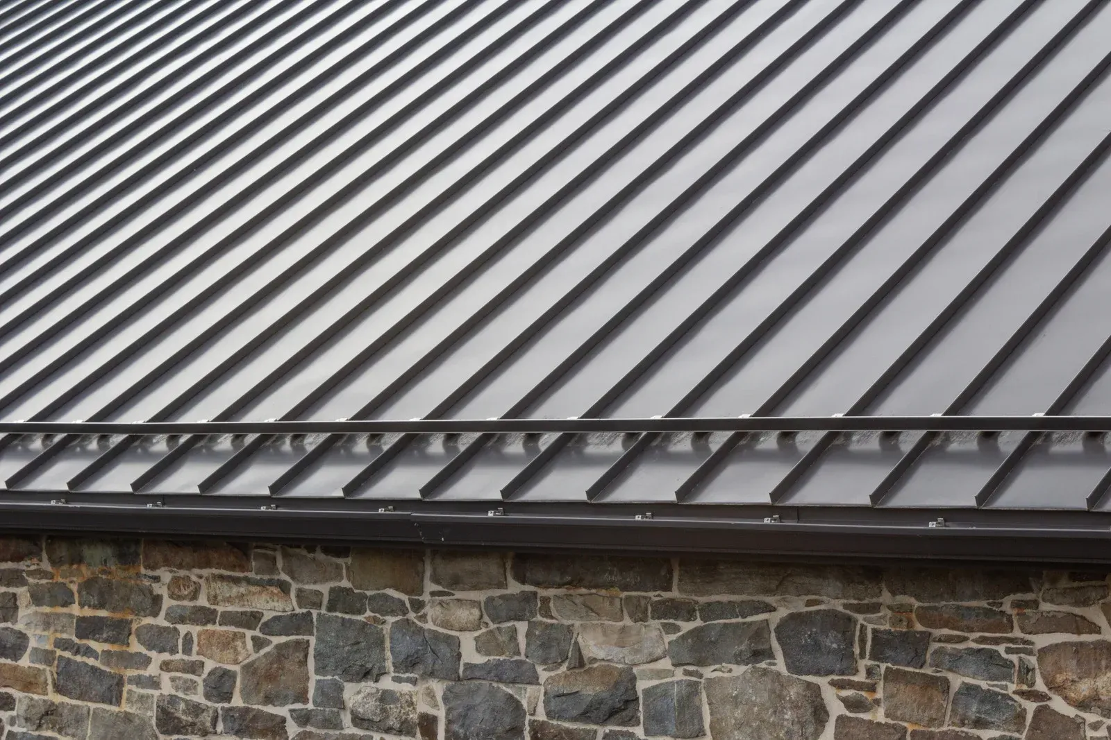 Gray metal roof with ribbed design over a stone wall; includes a dark gutter along the edge.