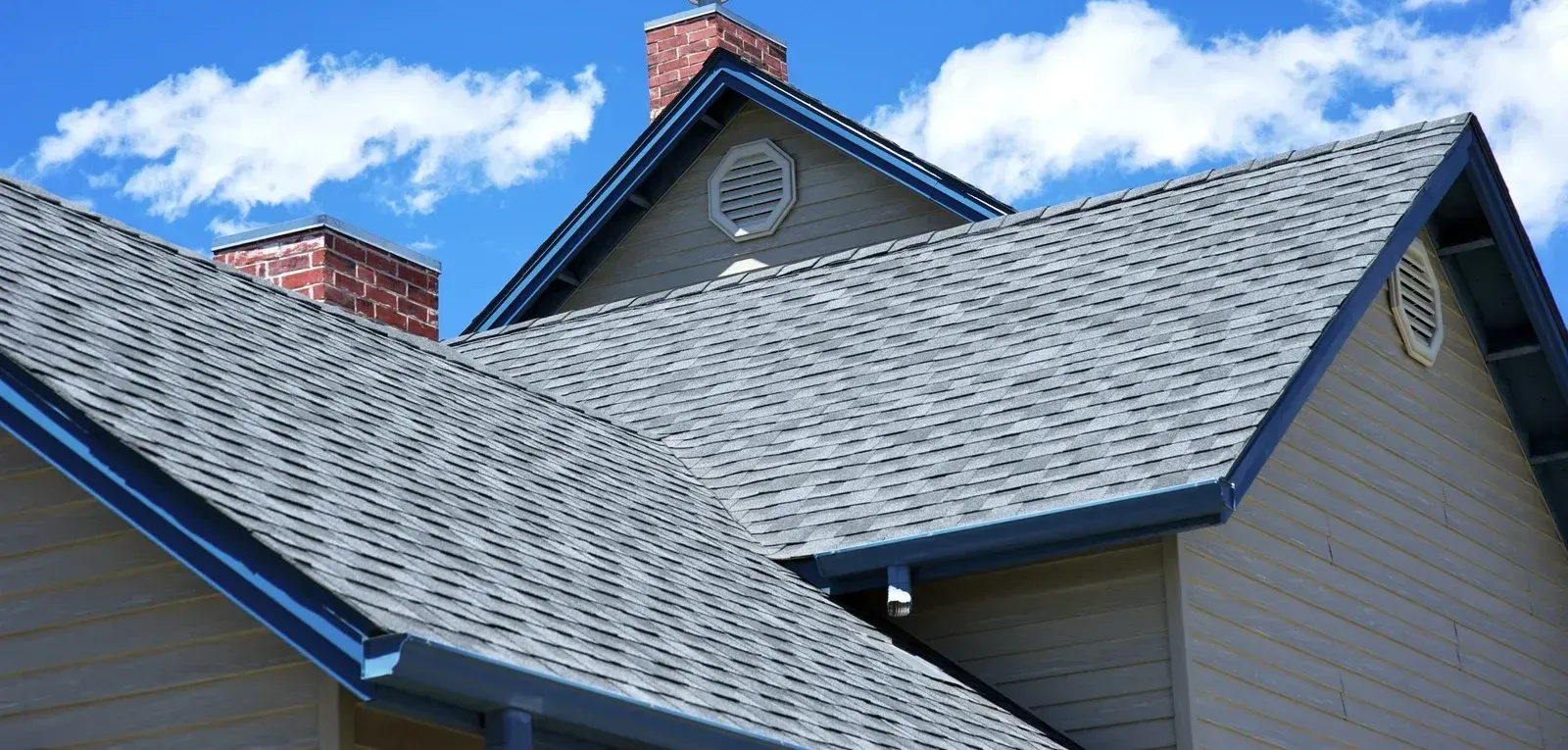 Roofs of a house against a bright blue sky with scattered white clouds. Two chimneys are visible.