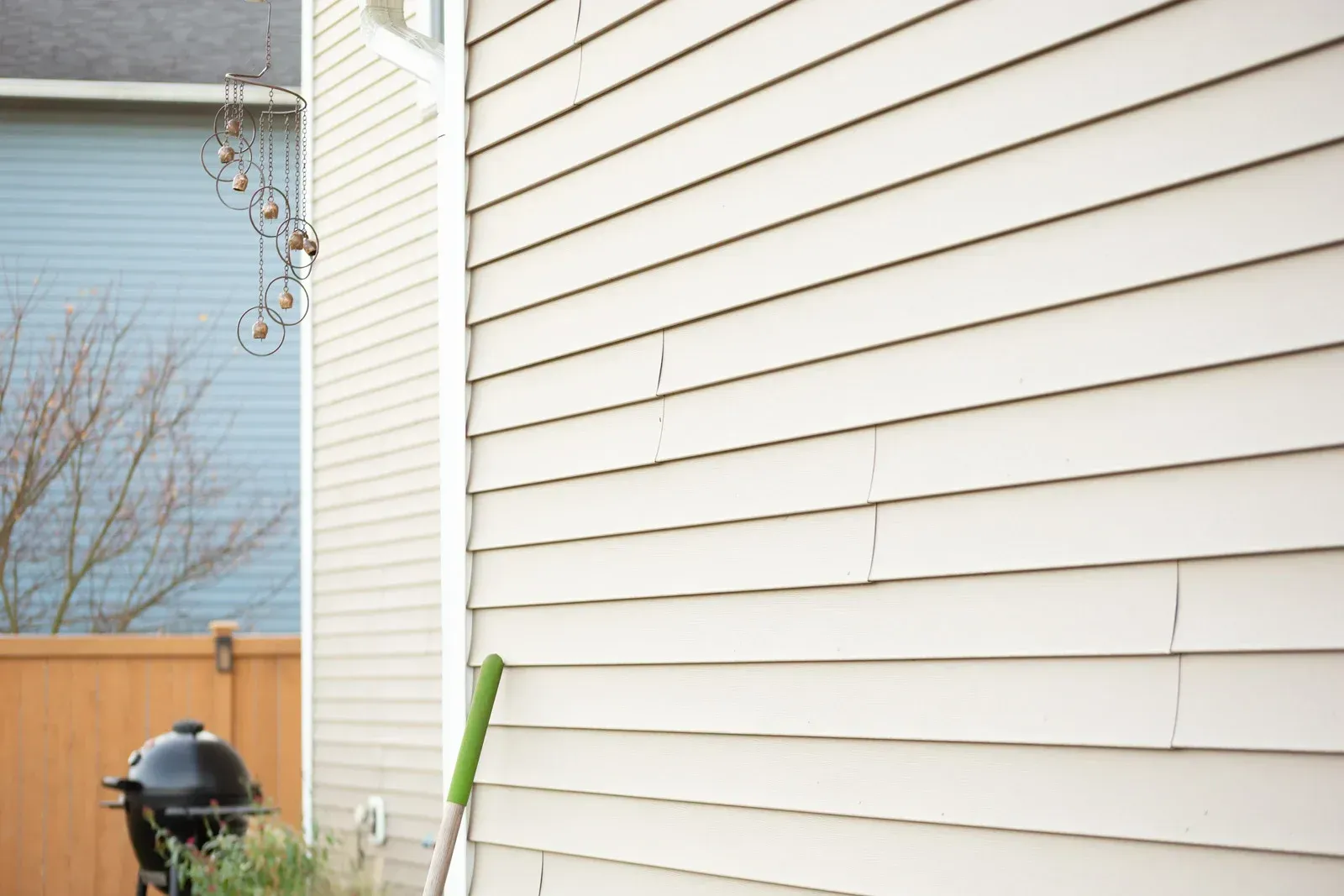 Beige siding of a house with visible cracks. In the background, a wooden fence, grill, and a blue house are visible.