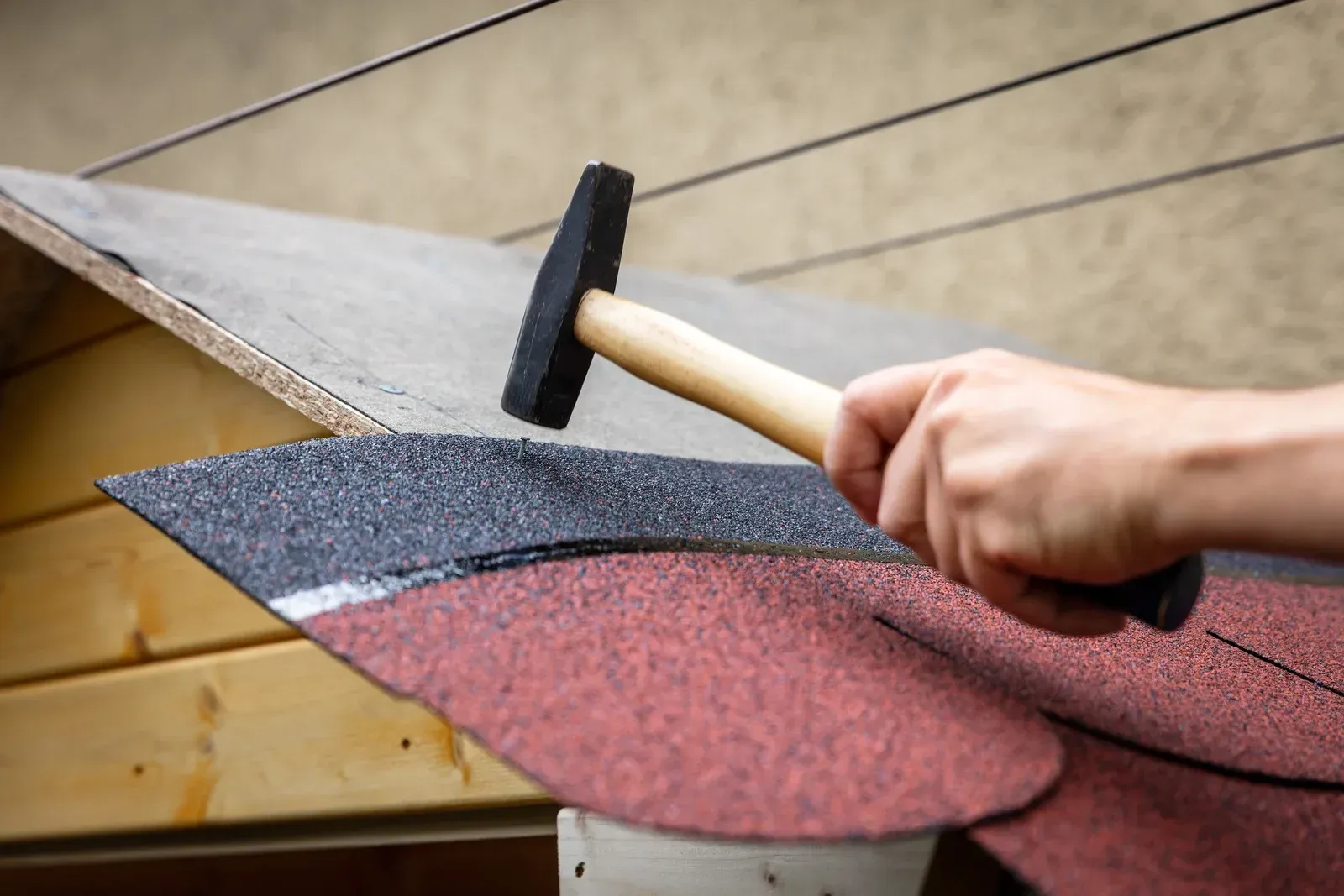 A person hammers a roofing shingle onto a wooden structure, with shingles in red and black.