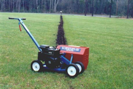 Lawn edger machine cutting a trench in green grass. Red casing, blue frame, black engine.