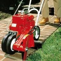 Red edging machine on a brick path, cutting grass. A person in tan pants stands nearby.