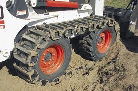 Close-up of a Bobcat skid-steer loader with track conversion kit. White machine, orange wheels, in a sandy setting.