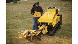 Man operating a yellow stump grinder on grass, wearing ear protection.