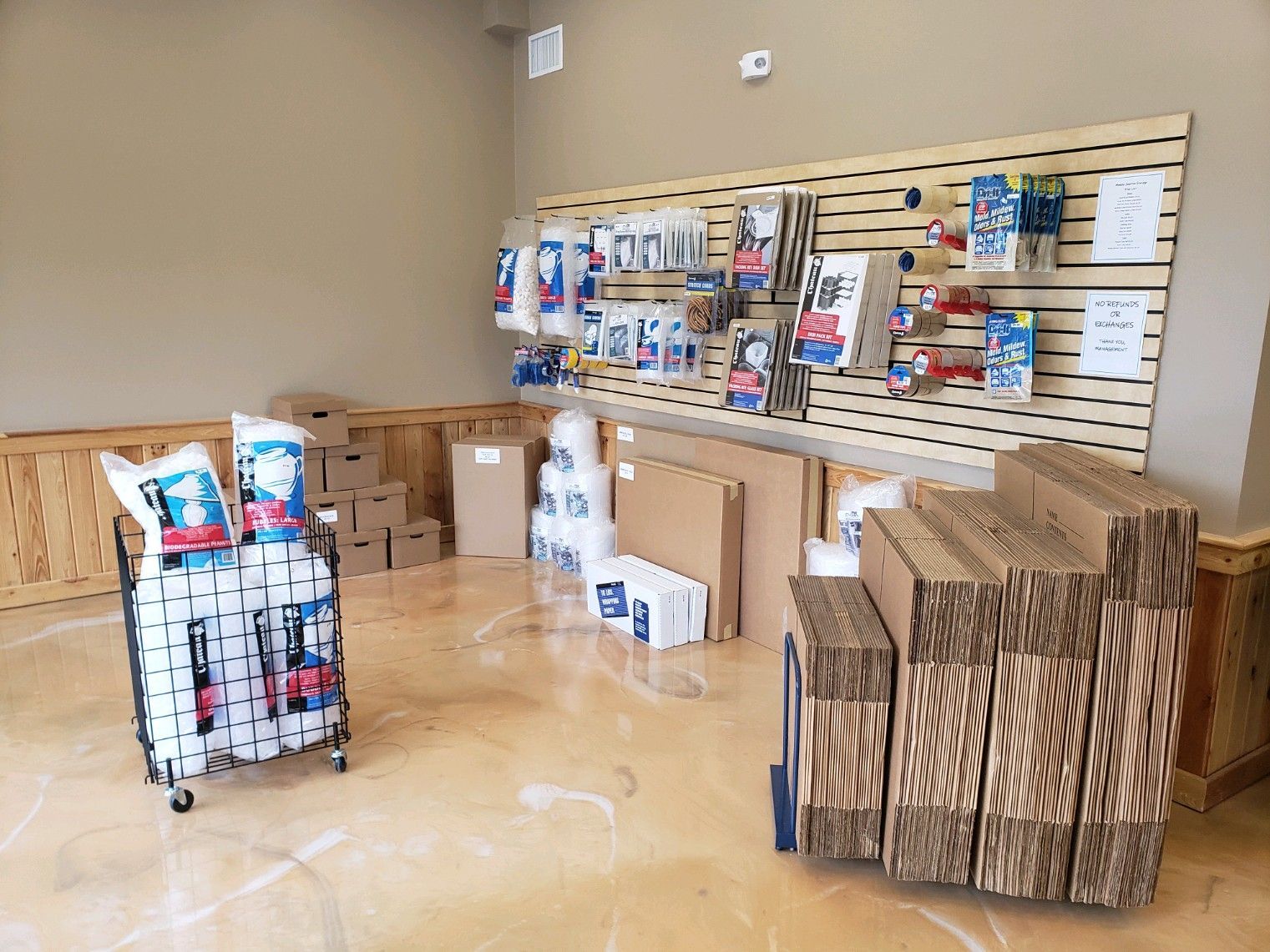 Storage supplies display inside a beige-walled store, including cardboard boxes, packing tape, bubble wrap.