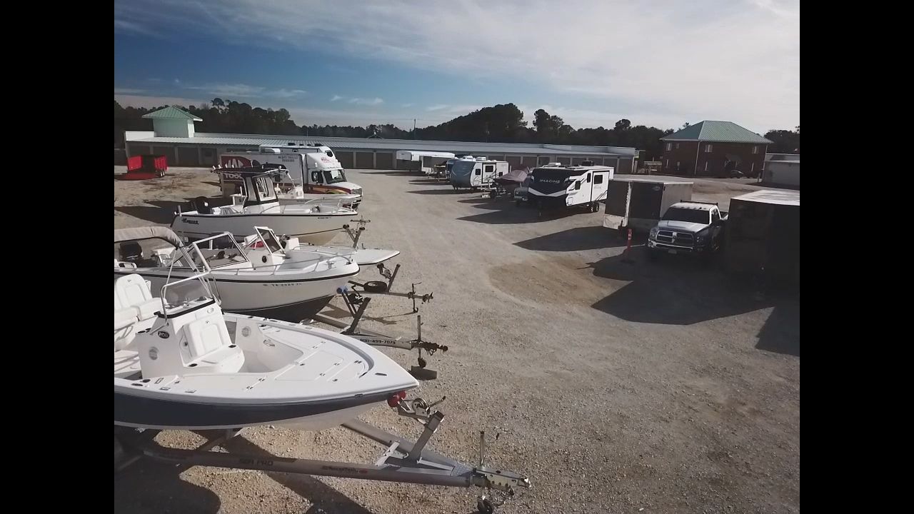 Boats and vehicles parked on a gravel lot near a body of water under a sunny sky.