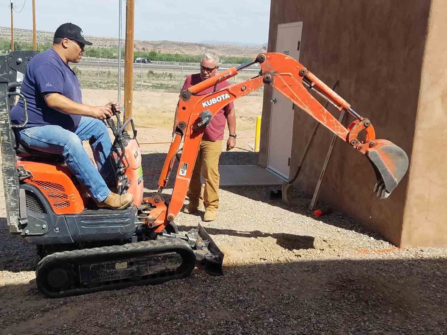 Two men are standing next to a small orange excavator.