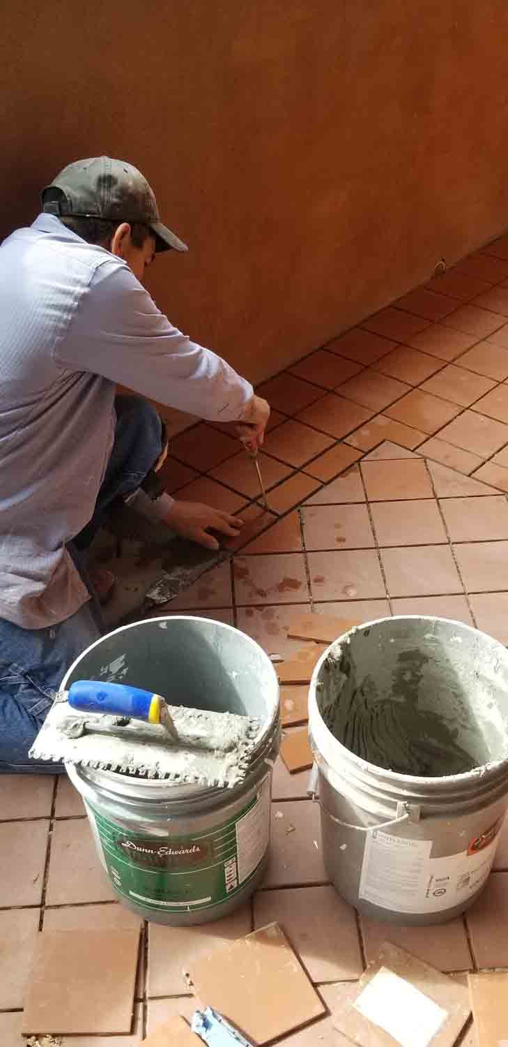 A man is kneeling down on a tiled floor next to two buckets of cement.