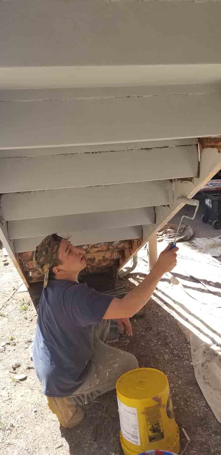 A man is kneeling down under a deck painting it.