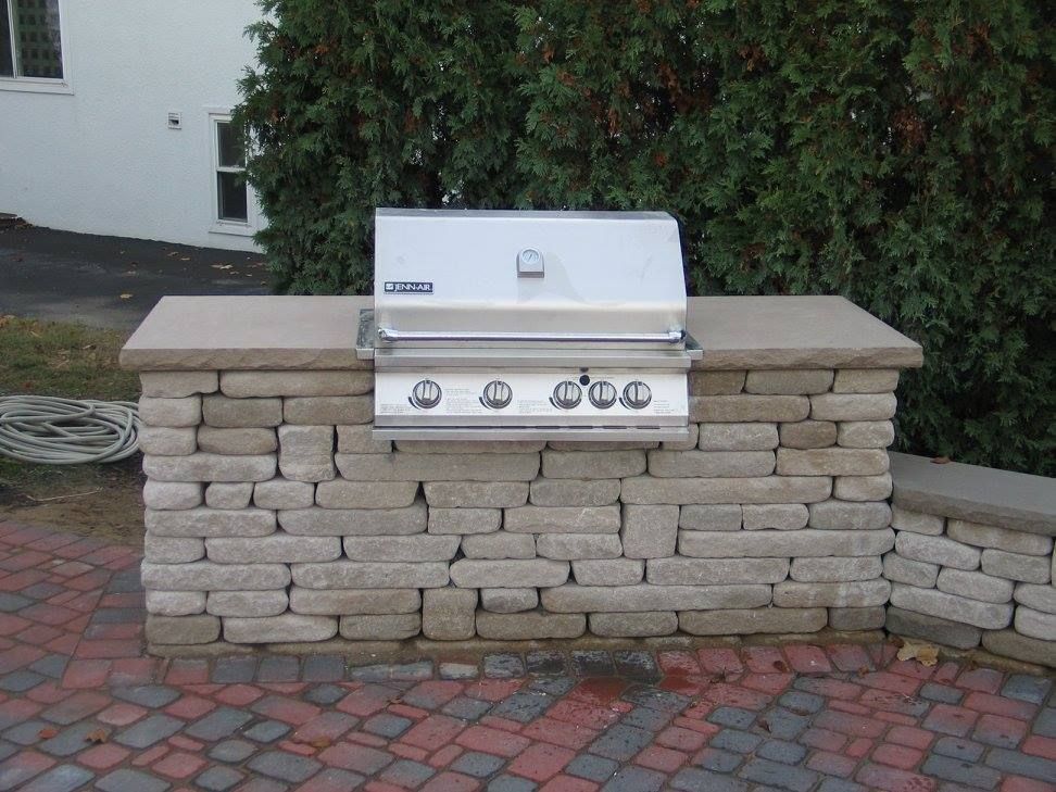 Outdoor built-in grill with stainless steel hood and front. Set in a stone brick base on a brick patio.
