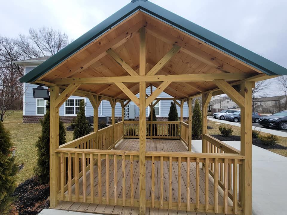 Wooden gazebo with green roof, deck, and railing, set outside with buildings and cars visible.