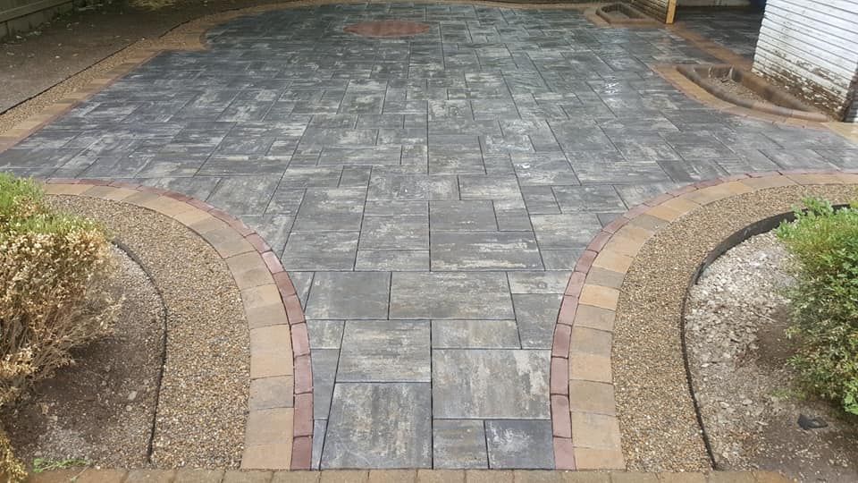 Stone patio with curved gravel and brick borders, edged by plants and a concrete walkway.